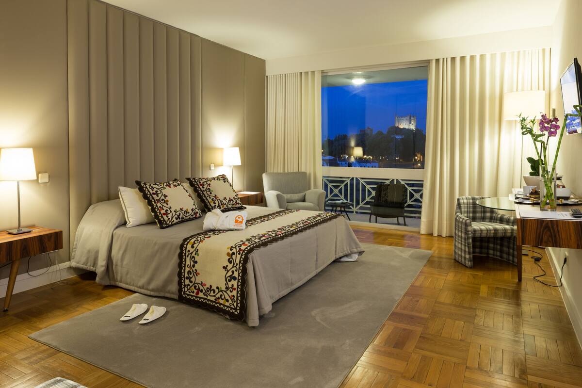 Ein Hotelzimmer mit einem großen Bett, einem Schreibtisch, einem Stuhl und Blick auf eine Burg bei Nacht.