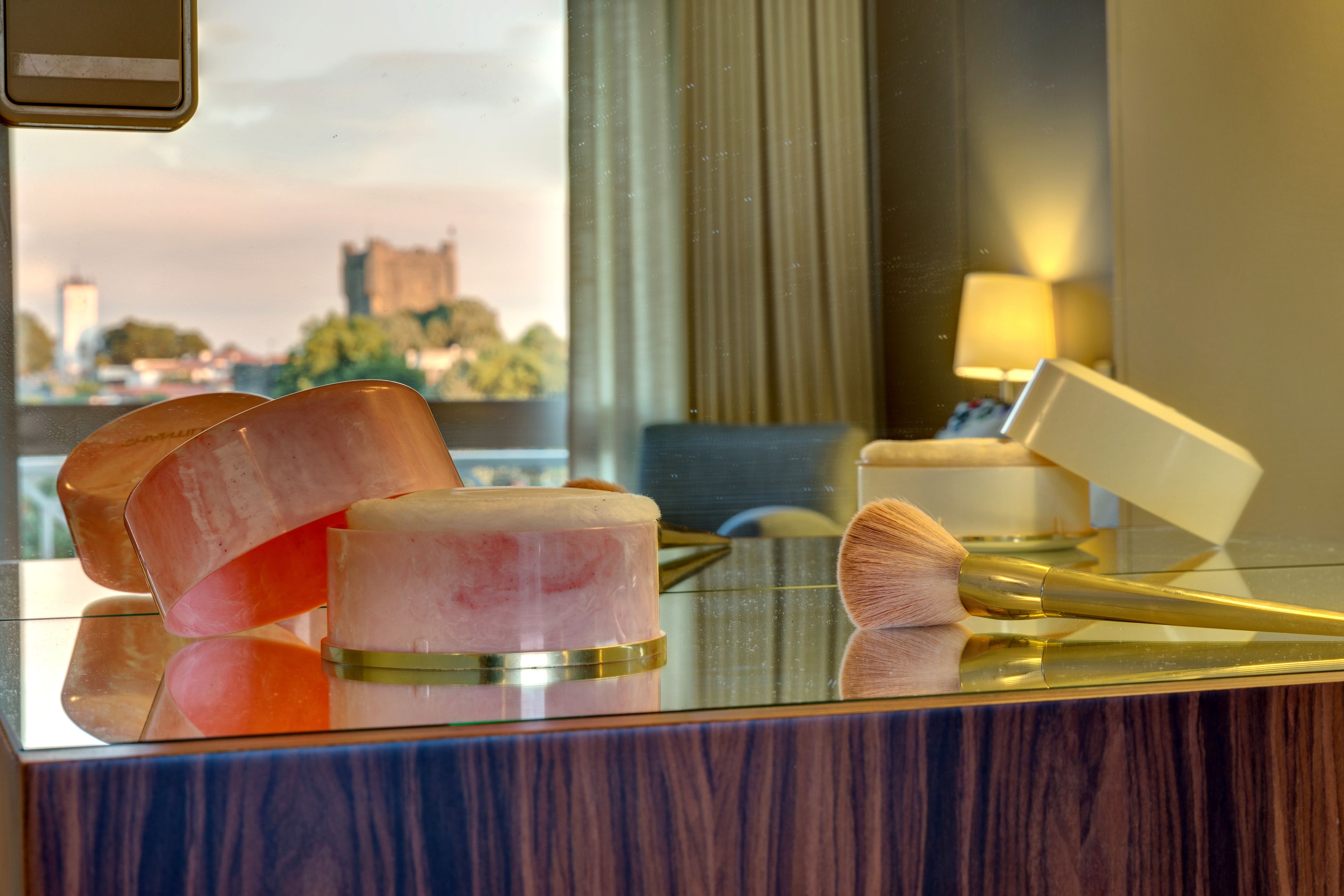 A hotel room with a glass table displaying makeup items and a reflection of a cityscape in the window.