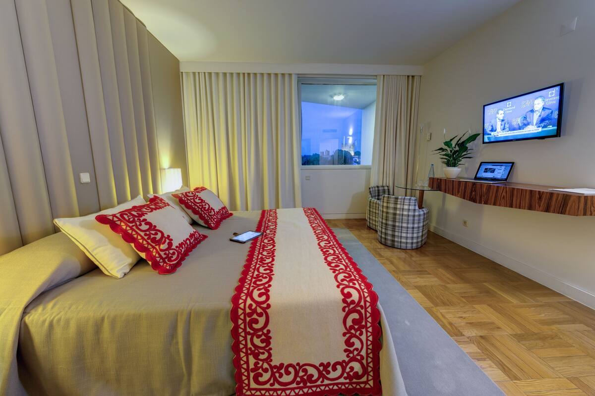 Ein Hotelzimmer mit Bett, Fernseher, einem Laptop auf einem Schreibtisch und Blick auf die Stadt durch das Fenster.