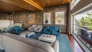 A modern lounge area with stone walls, wooden ceiling, and large windows offering a view of greenery. - Pousada de Bragança - São Bartolomeu (Braganca)