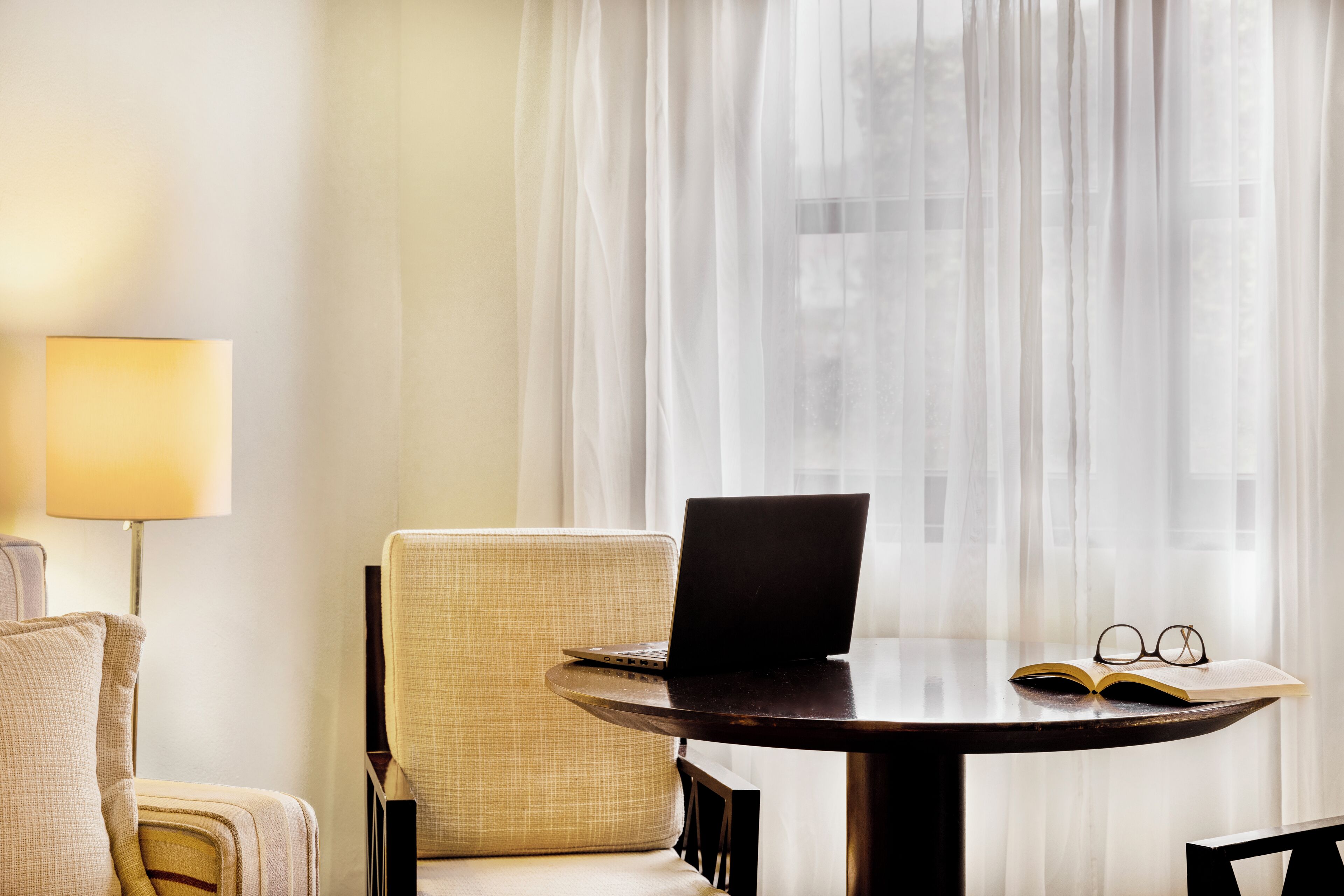 A room with a round table, a laptop, an open book, and a pair of glasses on the table.