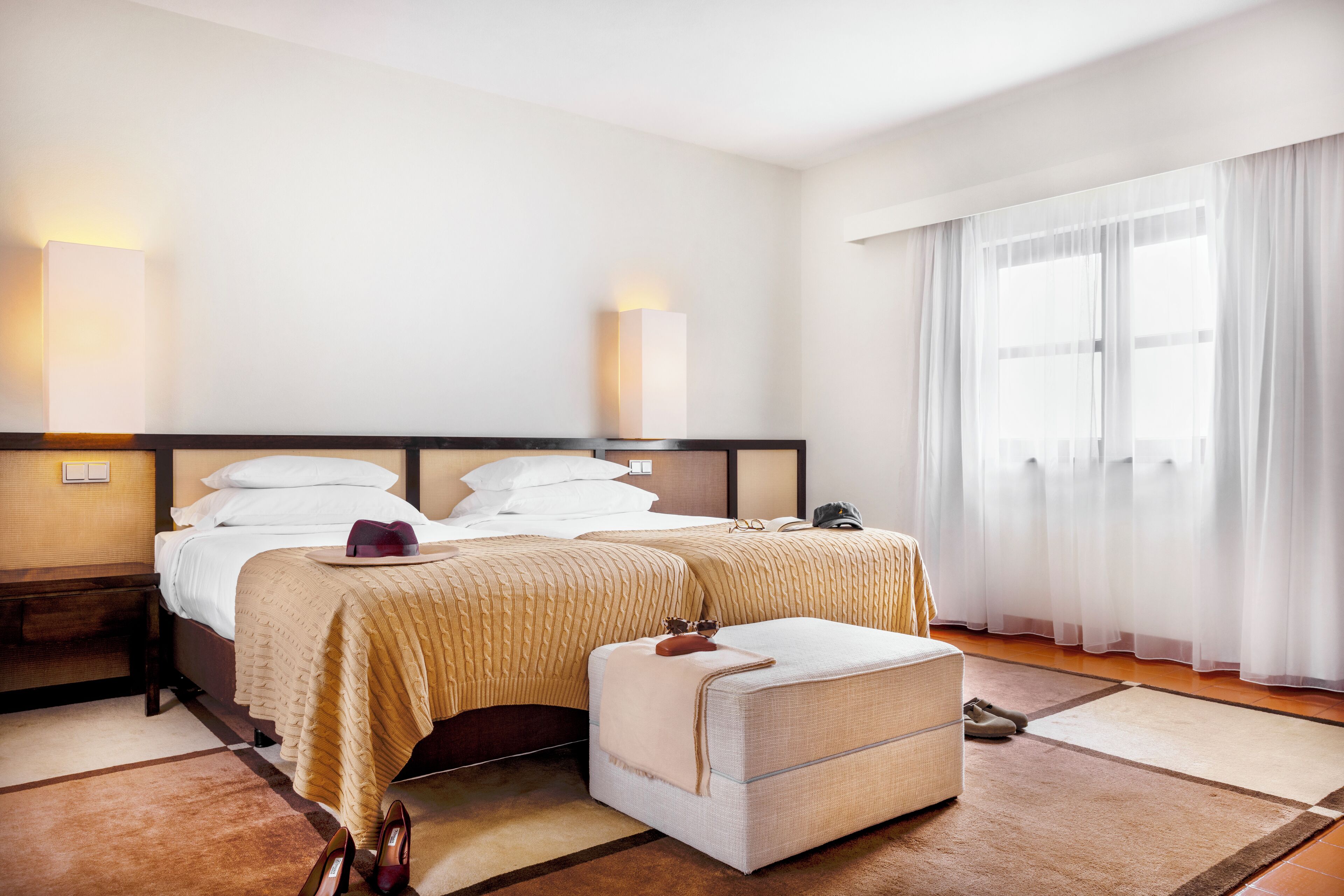 Habitación de hotel con cama doble, dos mesitas de noche, un banco y un ventanal con cortinas traslúcidas.