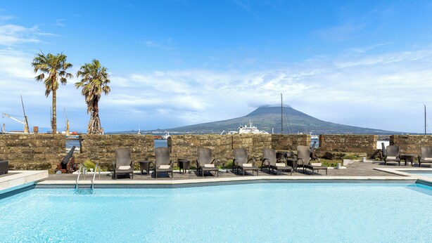 A pool with lounge chairs and palm trees, a stone wall, and a view of a volcano in the background. - Pousada Forte Horta (Horta)