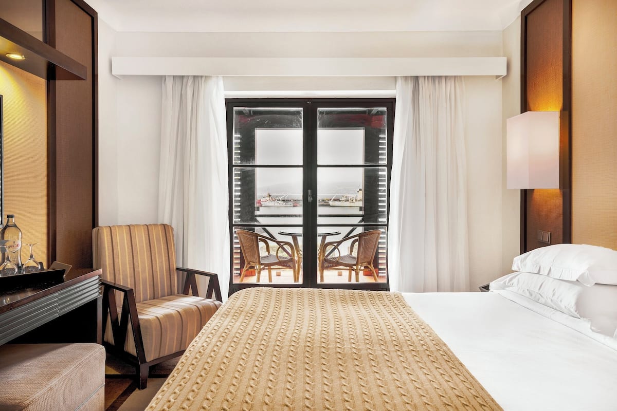 A hotel room with a bed, a chair, a desk, and a view of an outdoor balcony with tables and chairs.