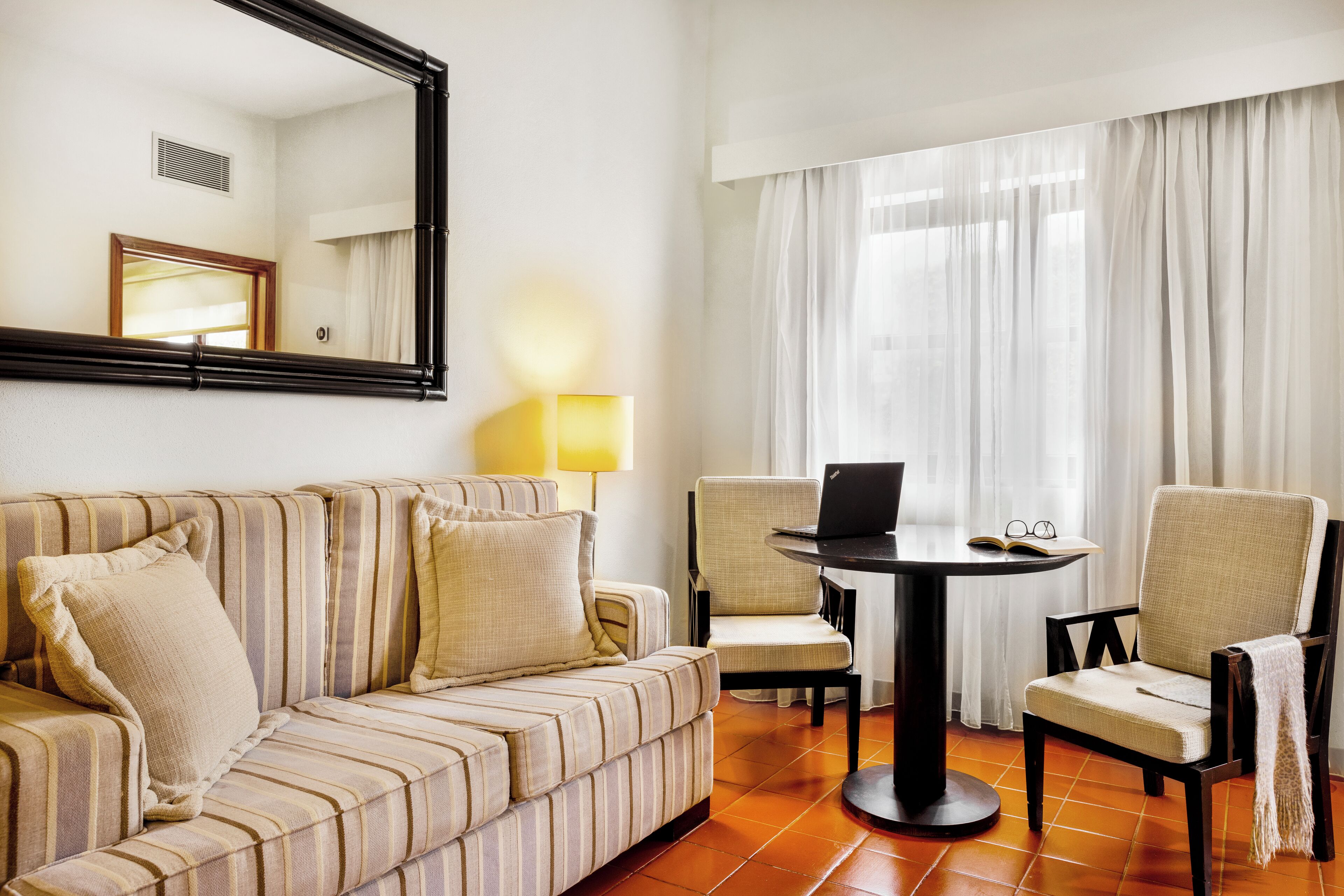Habitación de hotel con un sofá a rayas, dos sillones, una mesa redonda con una computadora portátil y un ventanal con cortinas.