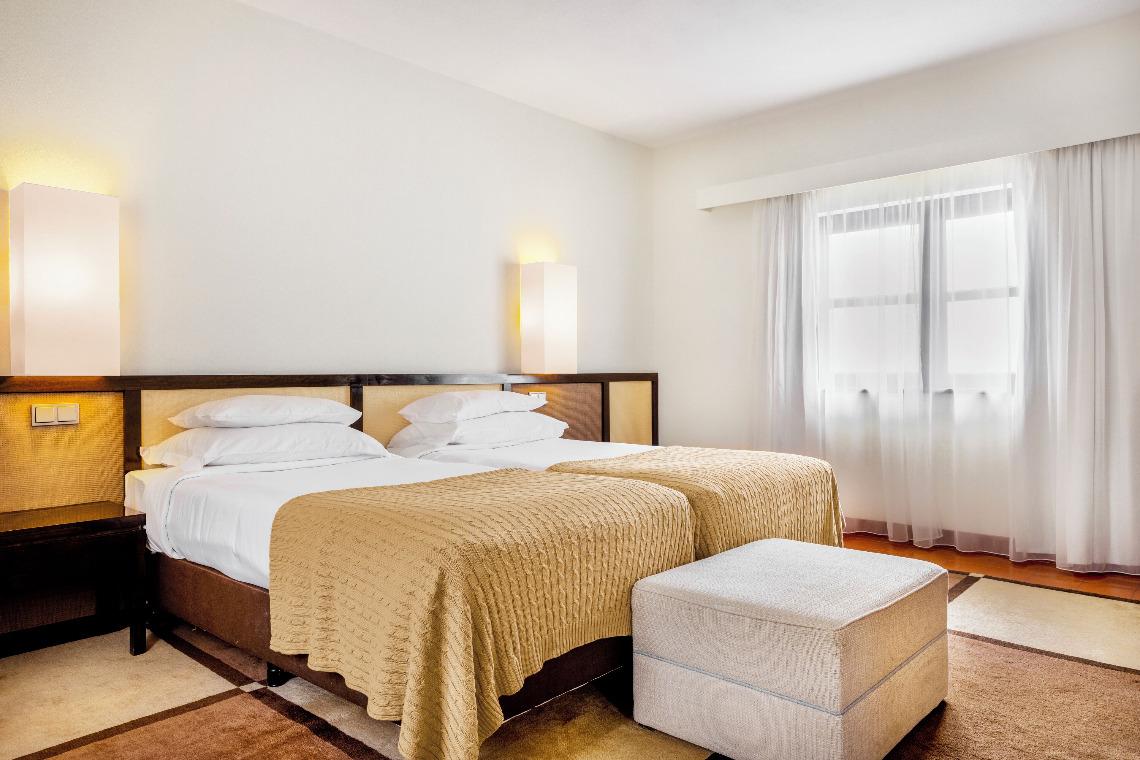 A hotel room with a double bed, bedside tables, a nightstand, a window with sheer curtains, and a view of the outside.