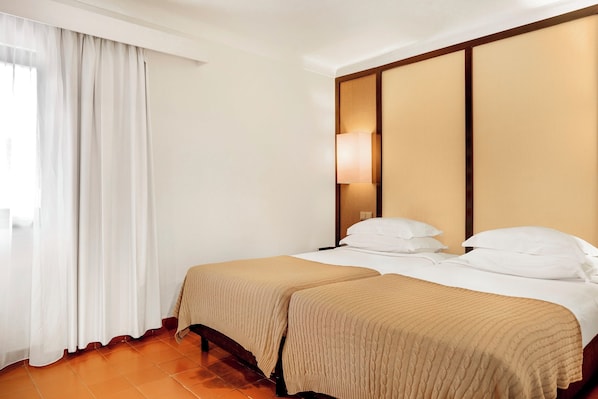 Une chambre d’hôtel avec deux lits, une grande fenêtre avec des rideaux blancs, une lampe fixée au mur et une tête de lit en bois.