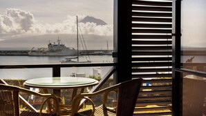 A balcony with wicker chairs and a glass table overlooking a harbor with a ship and a mountain in the background. - Pousada Forte Horta (Horta)