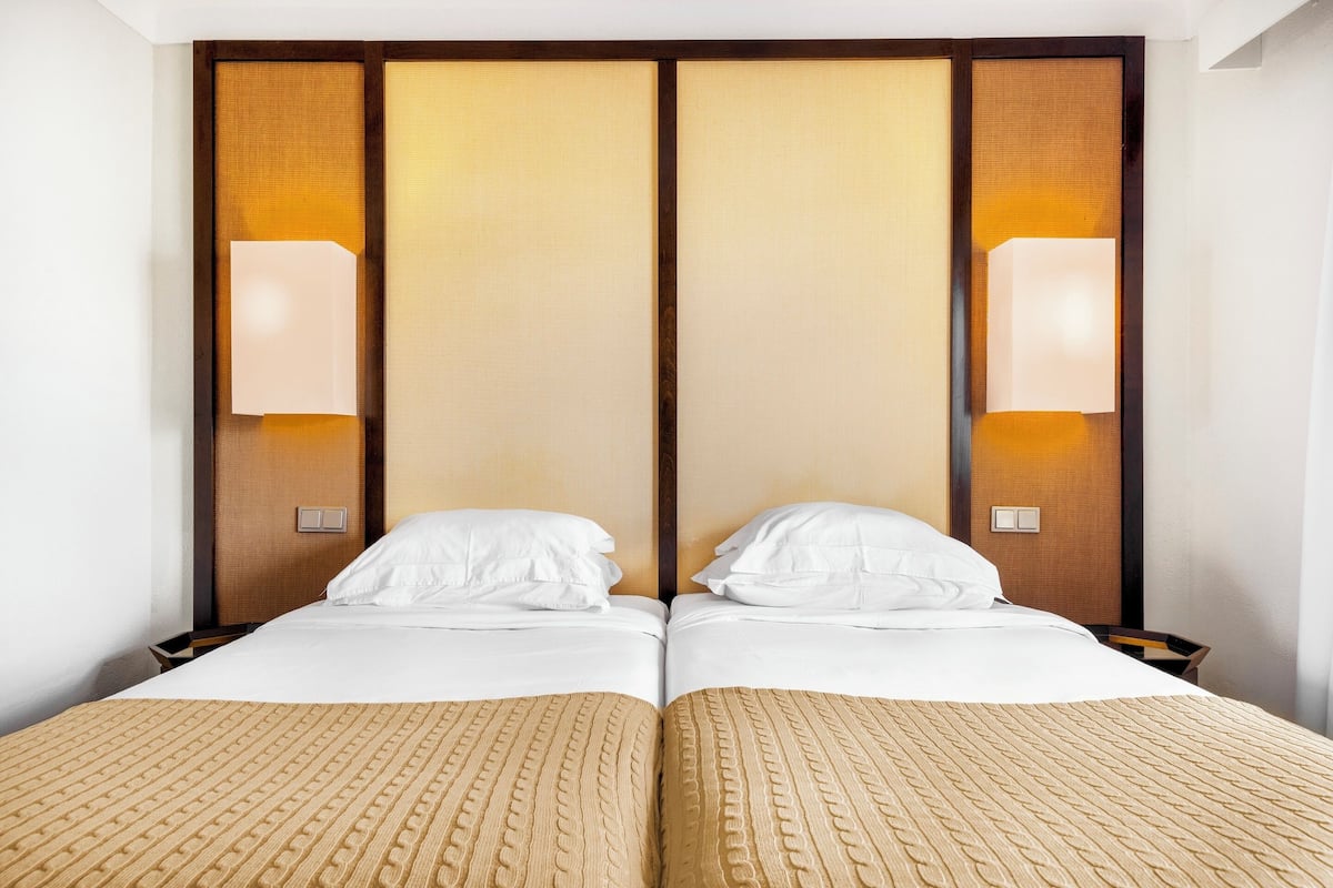 A double bed with a textured beige bedspread, two white pillows, and a wooden headboard with vertical panels and wall-mounted lamps.