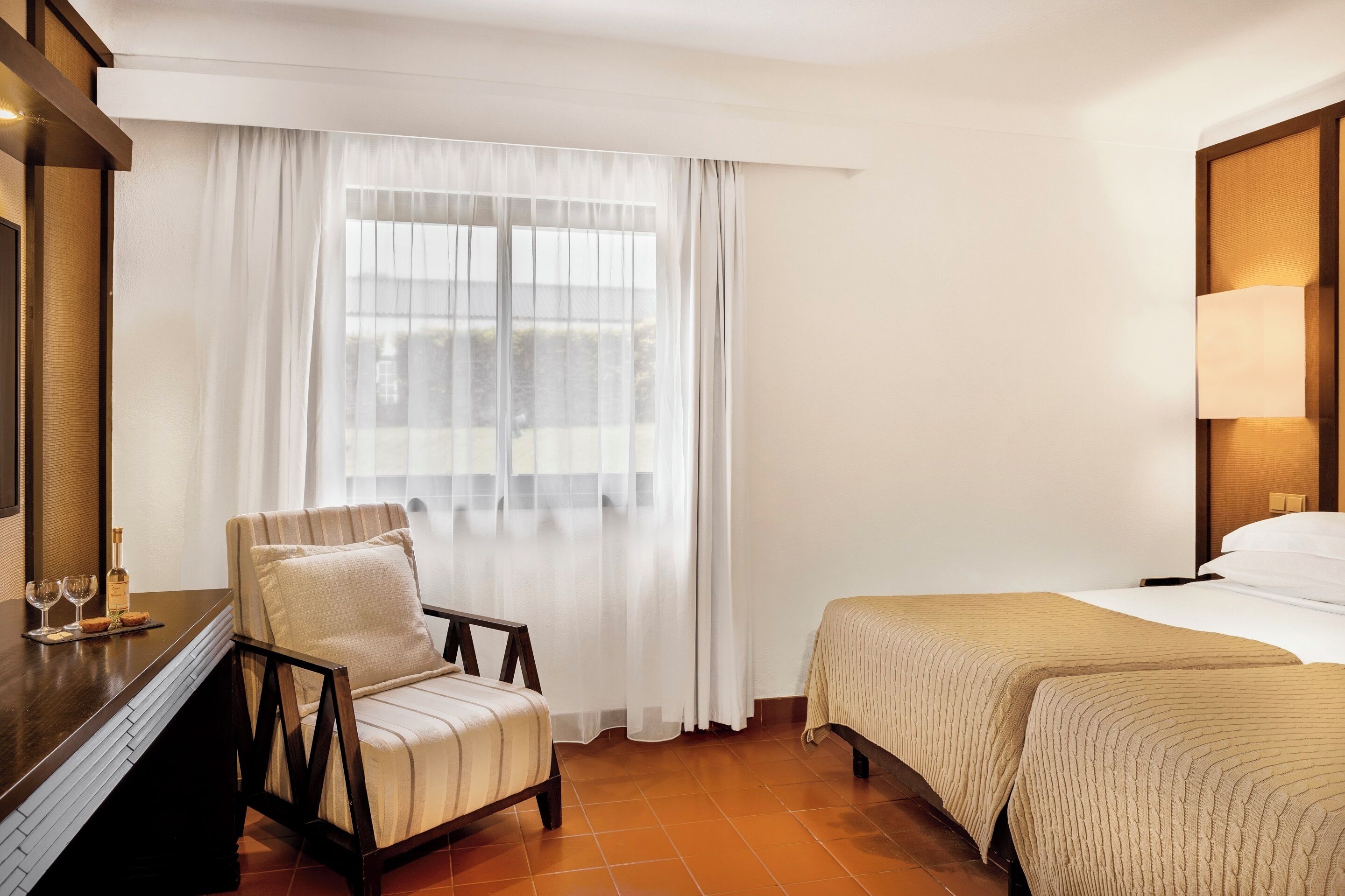 Une chambre d’hôtel avec un lit, une chaise, un bureau avec une lampe et une fenêtre avec des rideaux.
