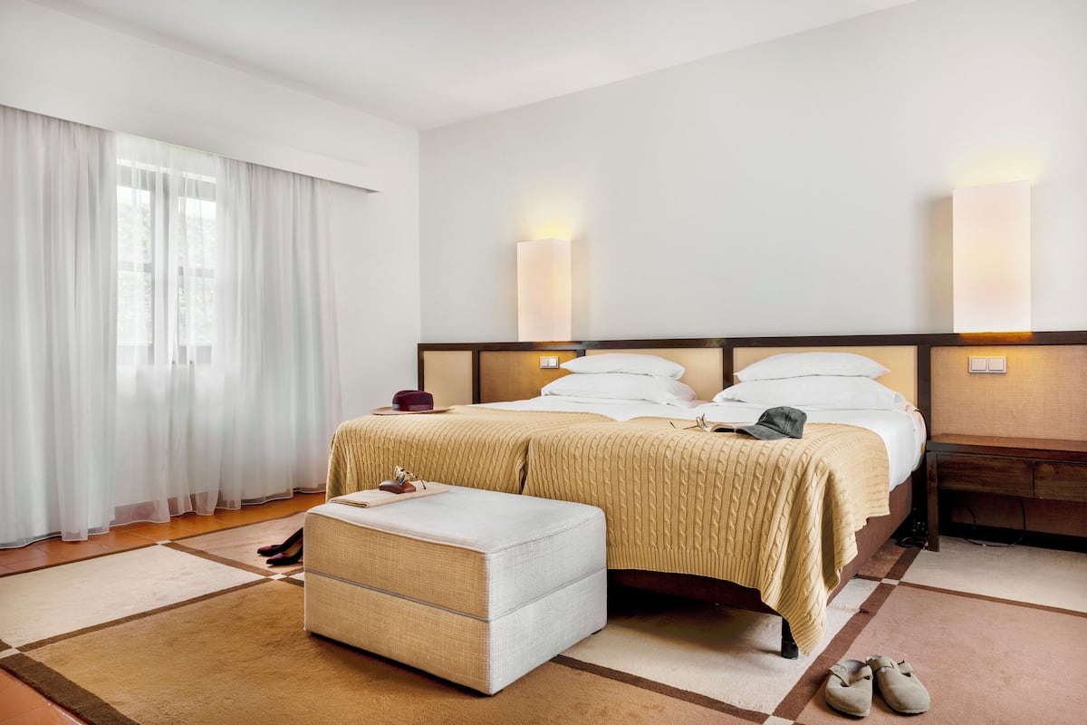 A hotel room with a bed, bedside tables, a nightstand, a window with curtains, and a pair of shoes on the floor.