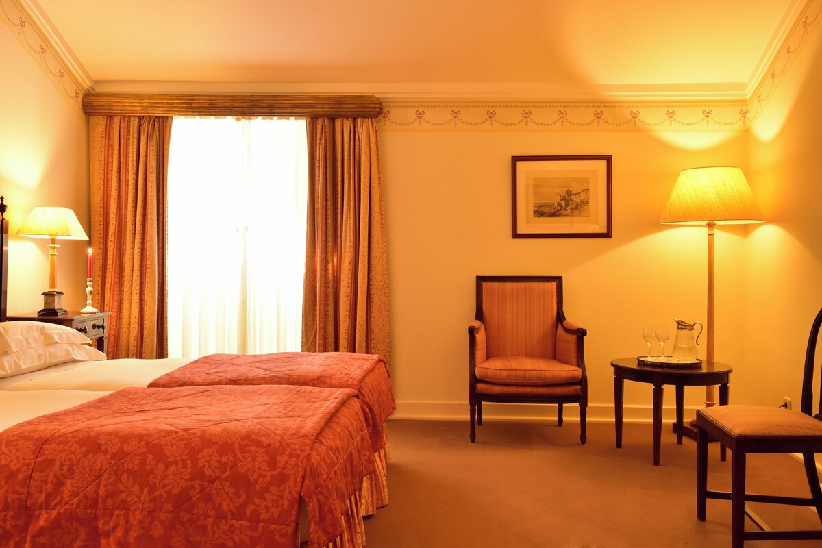A hotel room with a bed, two chairs, a table with a pitcher, and a framed picture on the wall.