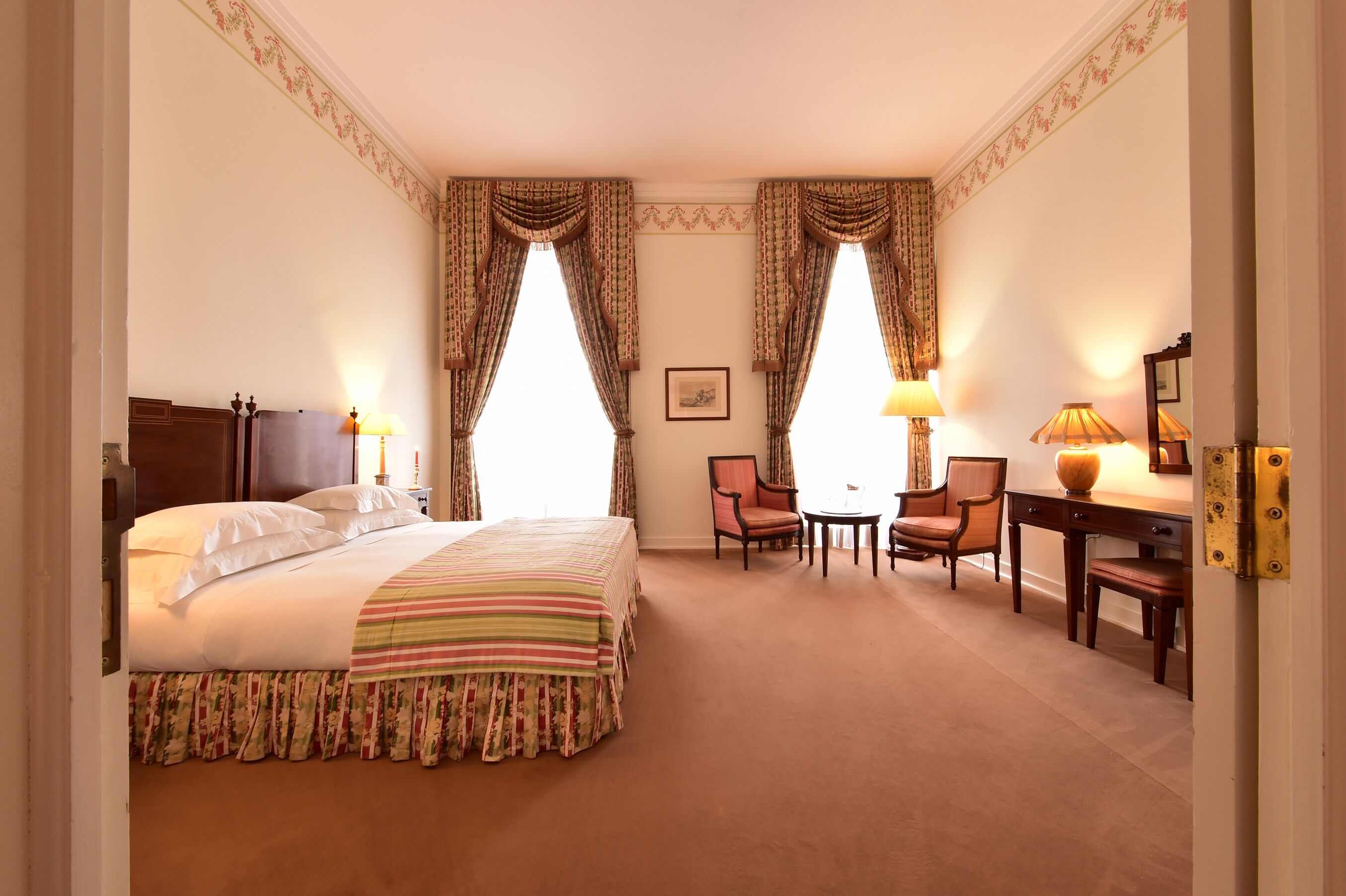 A hotel room with a large bed, two chairs, a small table, and a desk with a lamp.