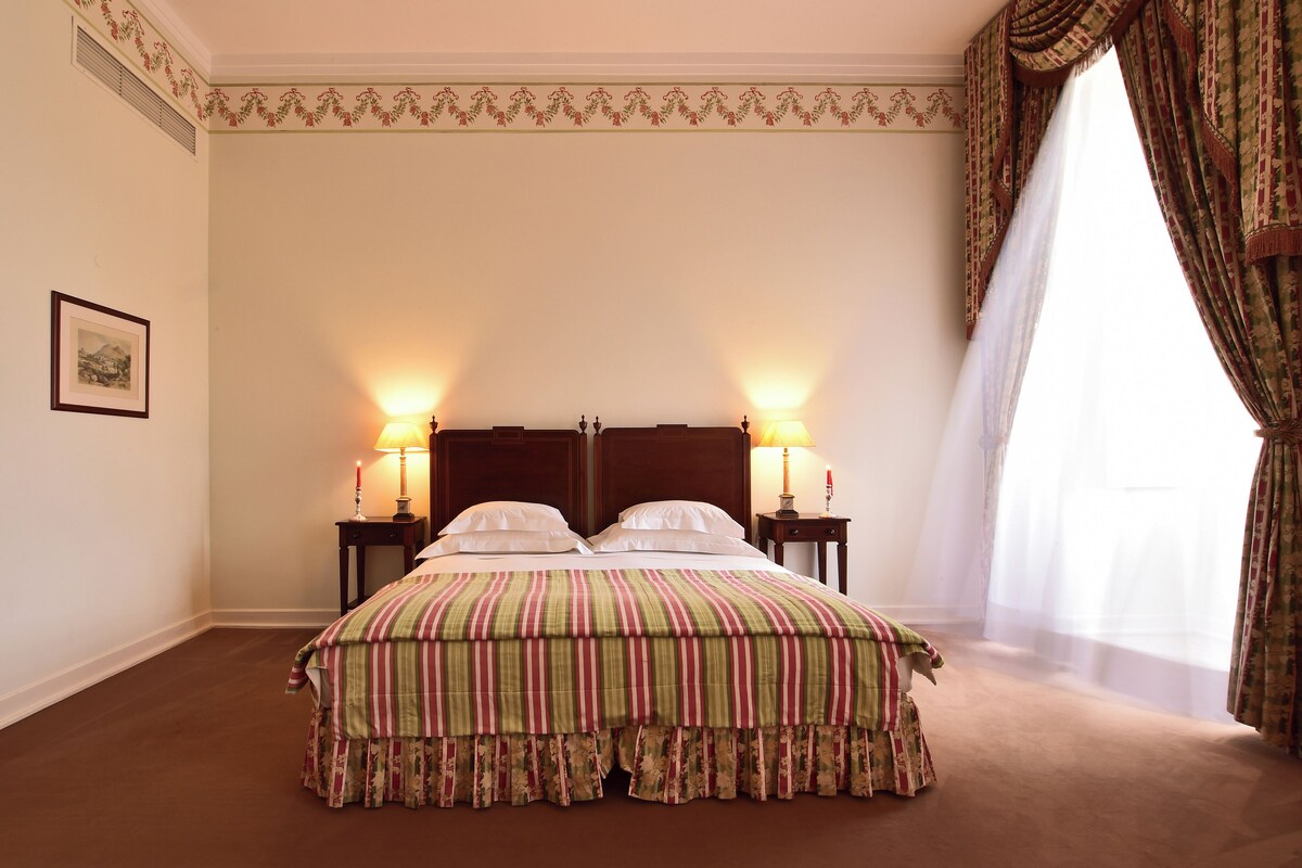 Habitación de hotel con una cama grande, dos mesitas de noche con lámparas, un cuadro enmarcado en la pared y un ventanal con cortinas.