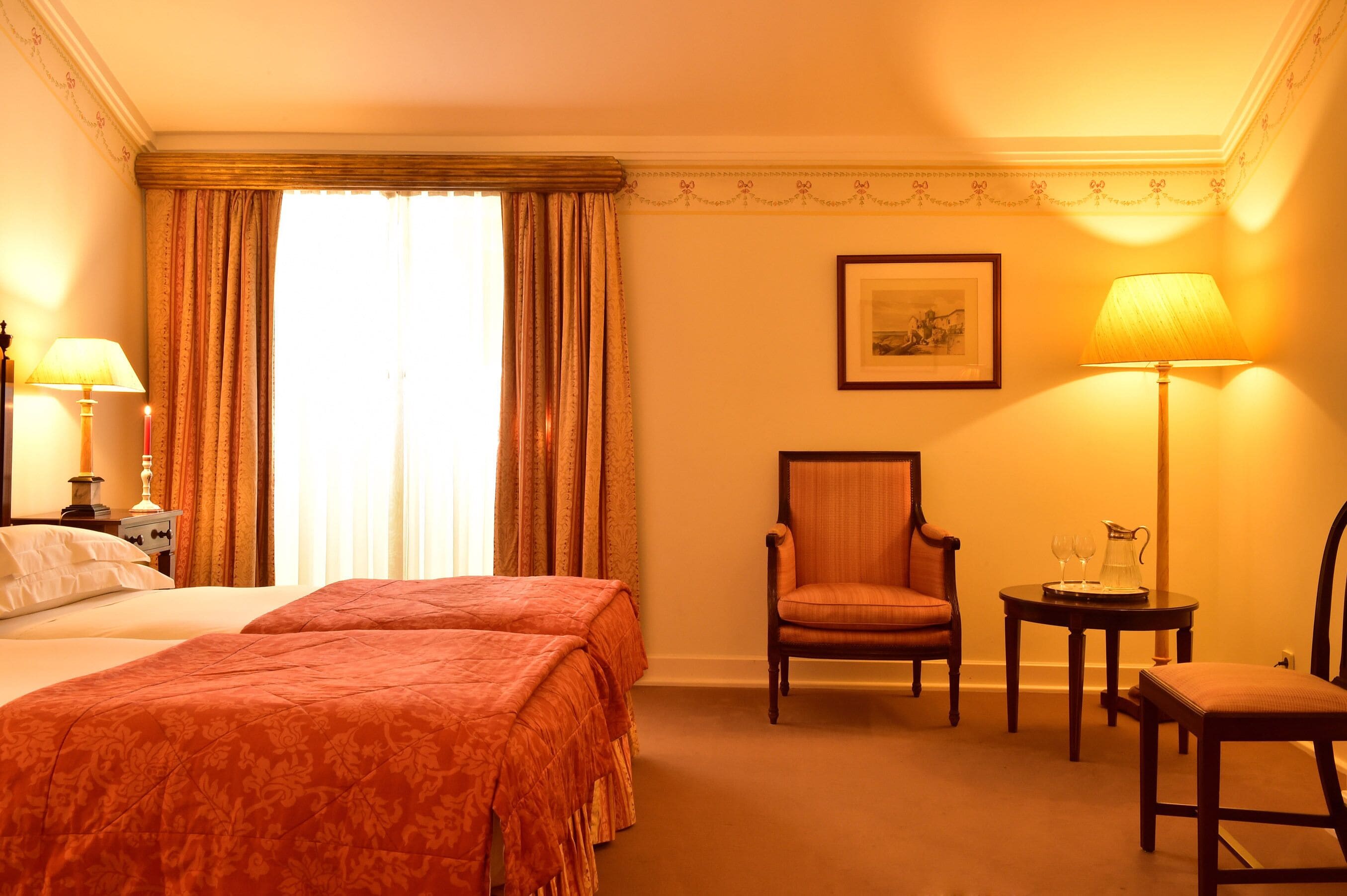 A hotel room with two beds, a chair, a table with a pitcher and glasses, and a framed picture on the wall.