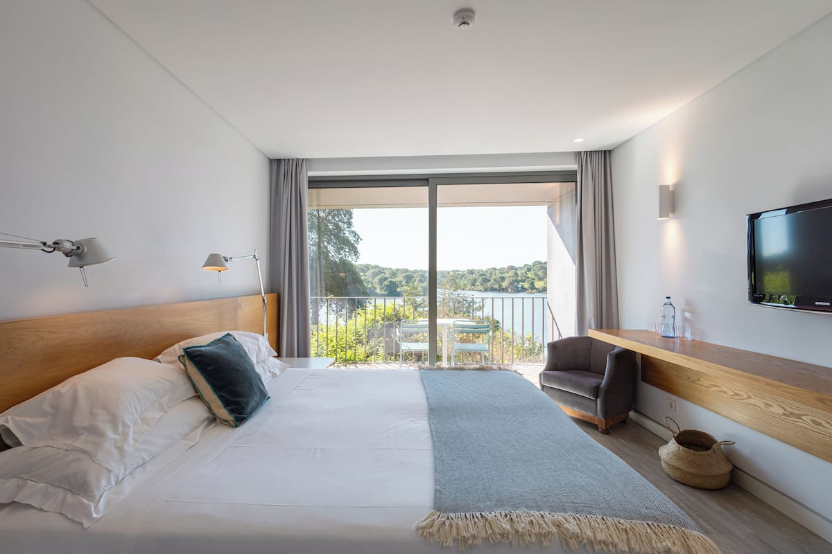 A modern hotel room with a large bed, a TV, a balcony with a view, and a wooden desk.