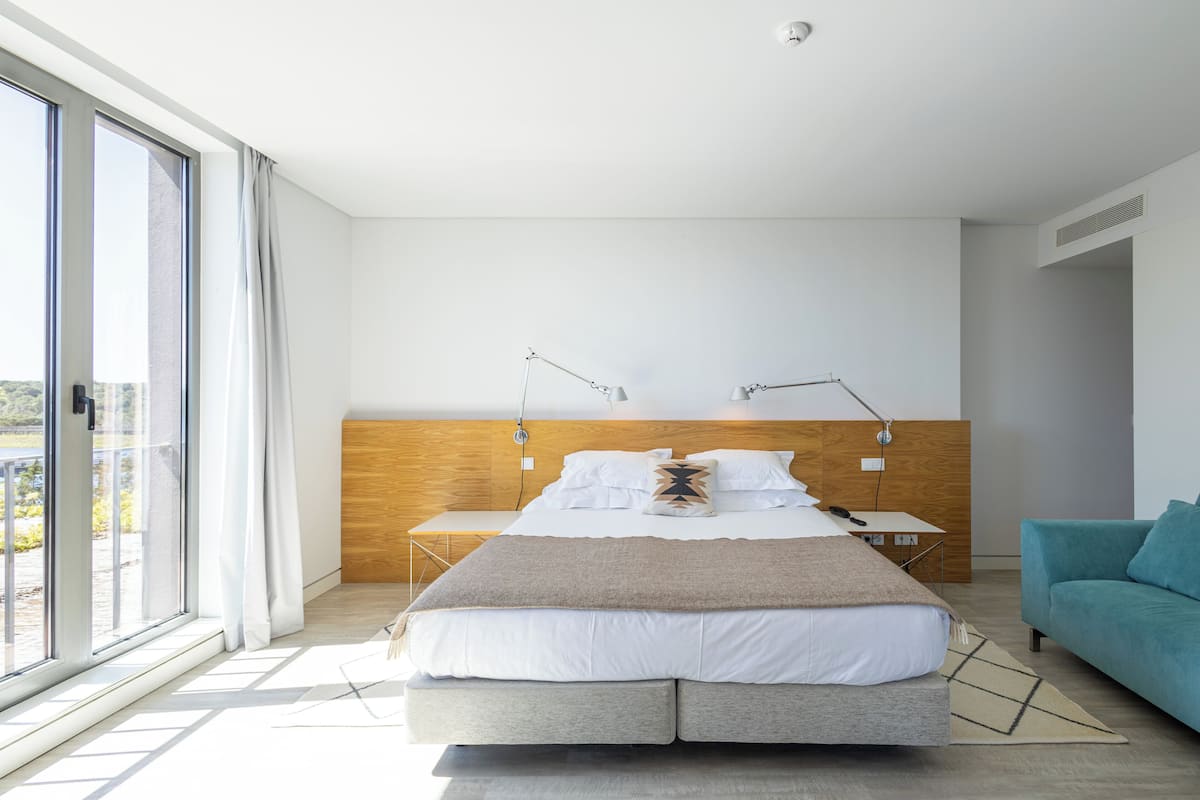 A modern bedroom with a large bed, bedside tables, a nightstand, a sofa, and a view through a large window.