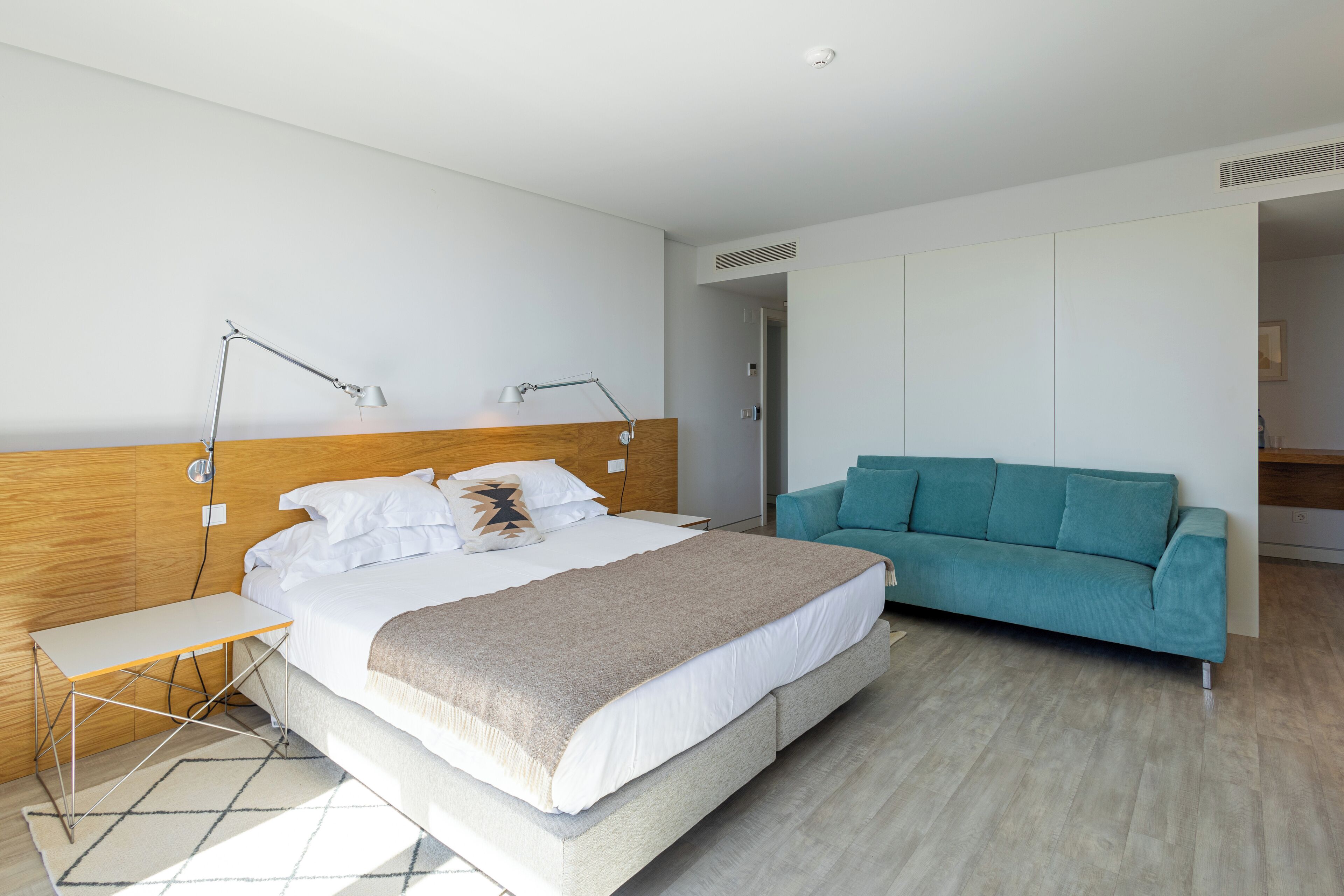 Ett modernt sovrum med en stor säng, ett sängbord, ett skrivbord, en turkosfärgad soffa och en sänggavel i trä.