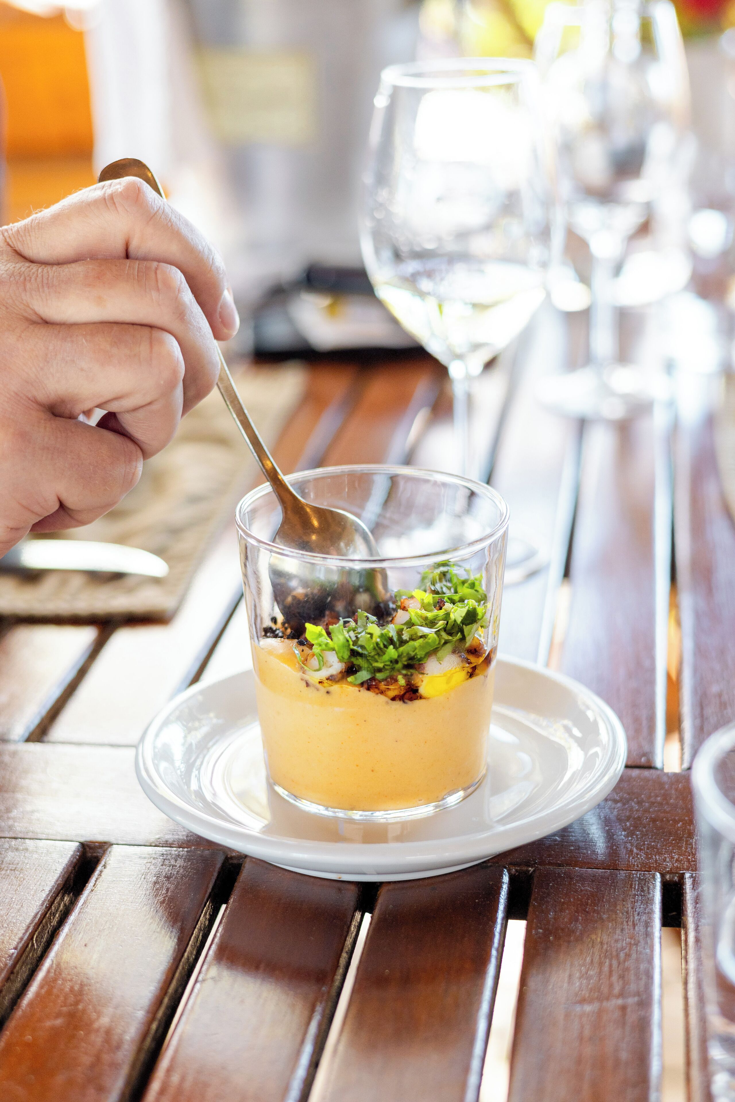 A dessert in a clear glass on a white plate, with a spoon in a hand, on a wooden table.