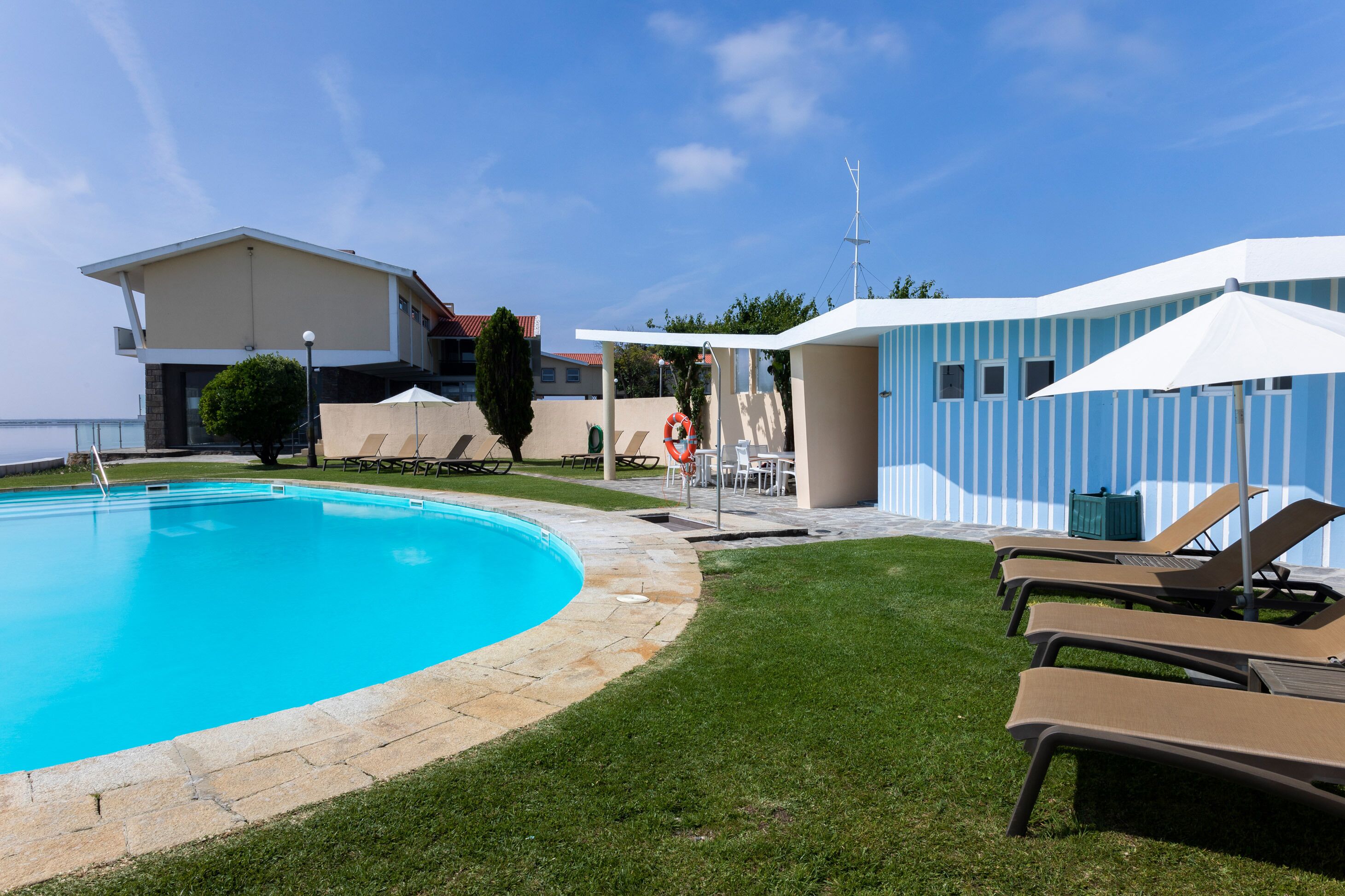 En swimmingpool med solstolar och parasoller, en byggnad med ett sluttande tak och en klar himmel.