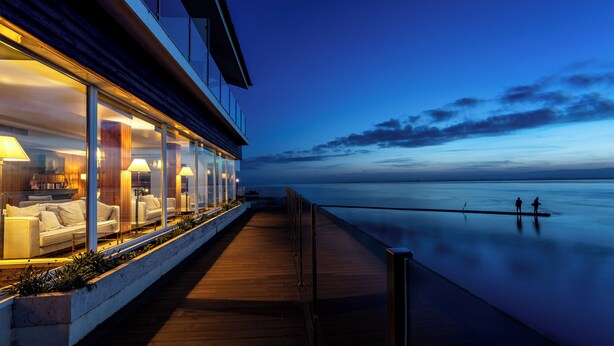 A waterfront hotel with a glass-enclosed lounge area, wooden deck, and a view of the calm sea.
