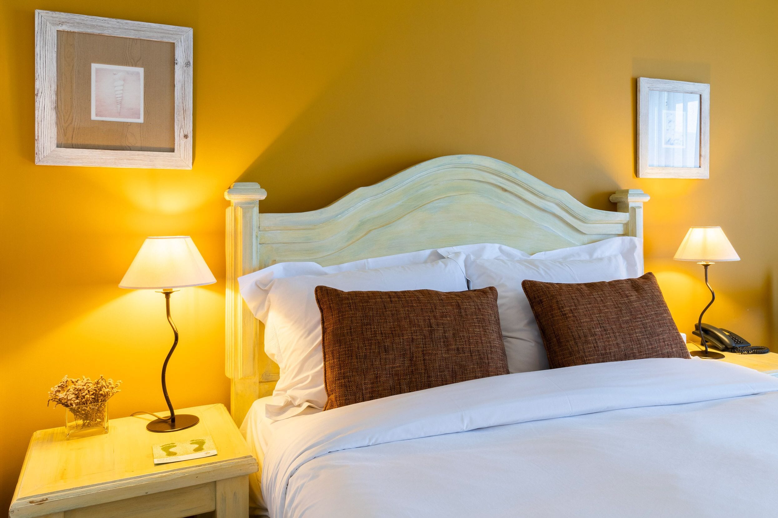 Un lit bien fait, avec du linge de lit blanc et deux oreillers marron, de part et d’autre duquel se trouvent deux tables de chevet avec des lampes et un téléphone.