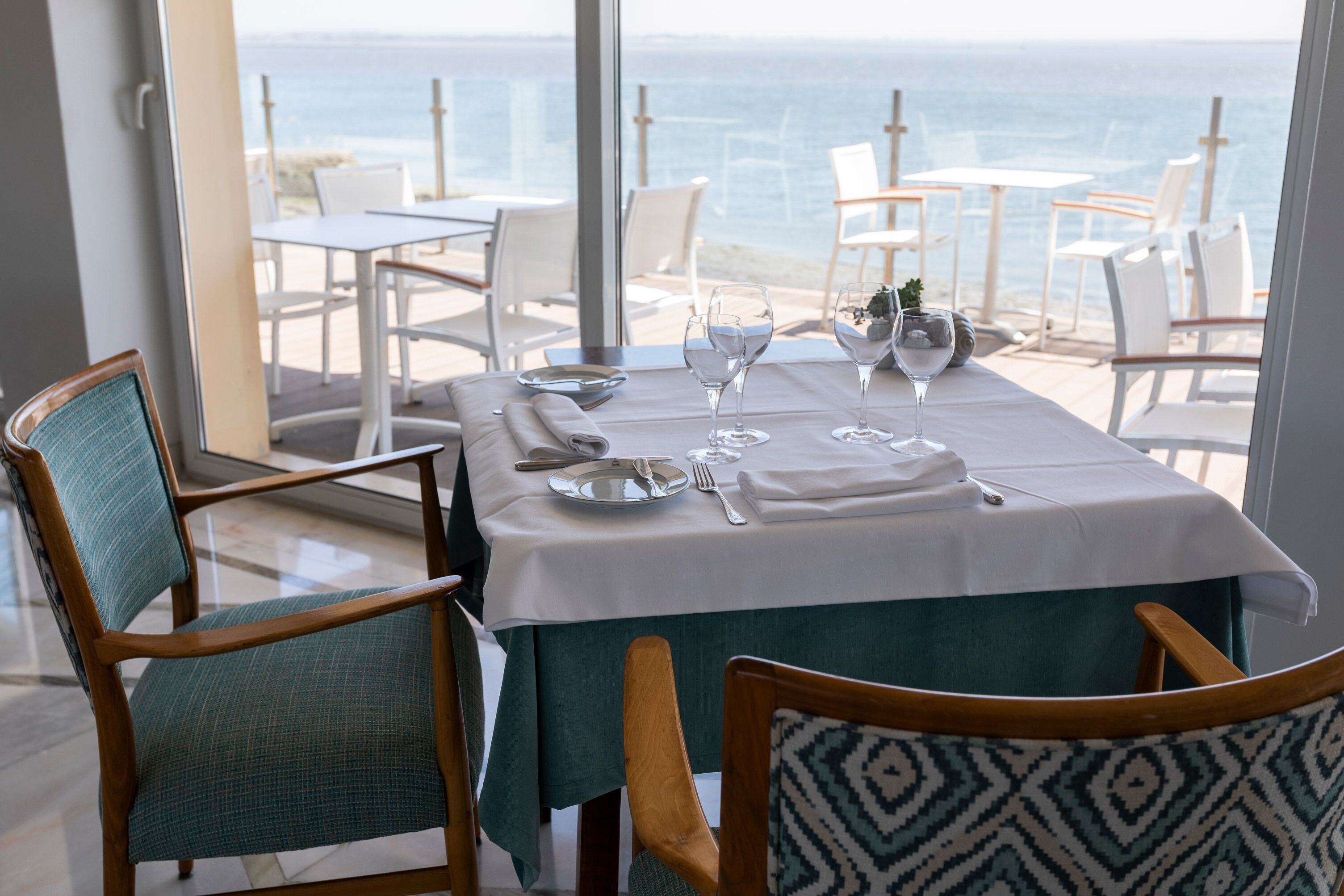 Een eettafel met uitzicht op de oceaan, witte stoelen en een blauwgroene tafelkleed.