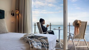 A hotel room with a bed, a balcony with a view of the sea, a table with a chair, and a person sitting at the table.