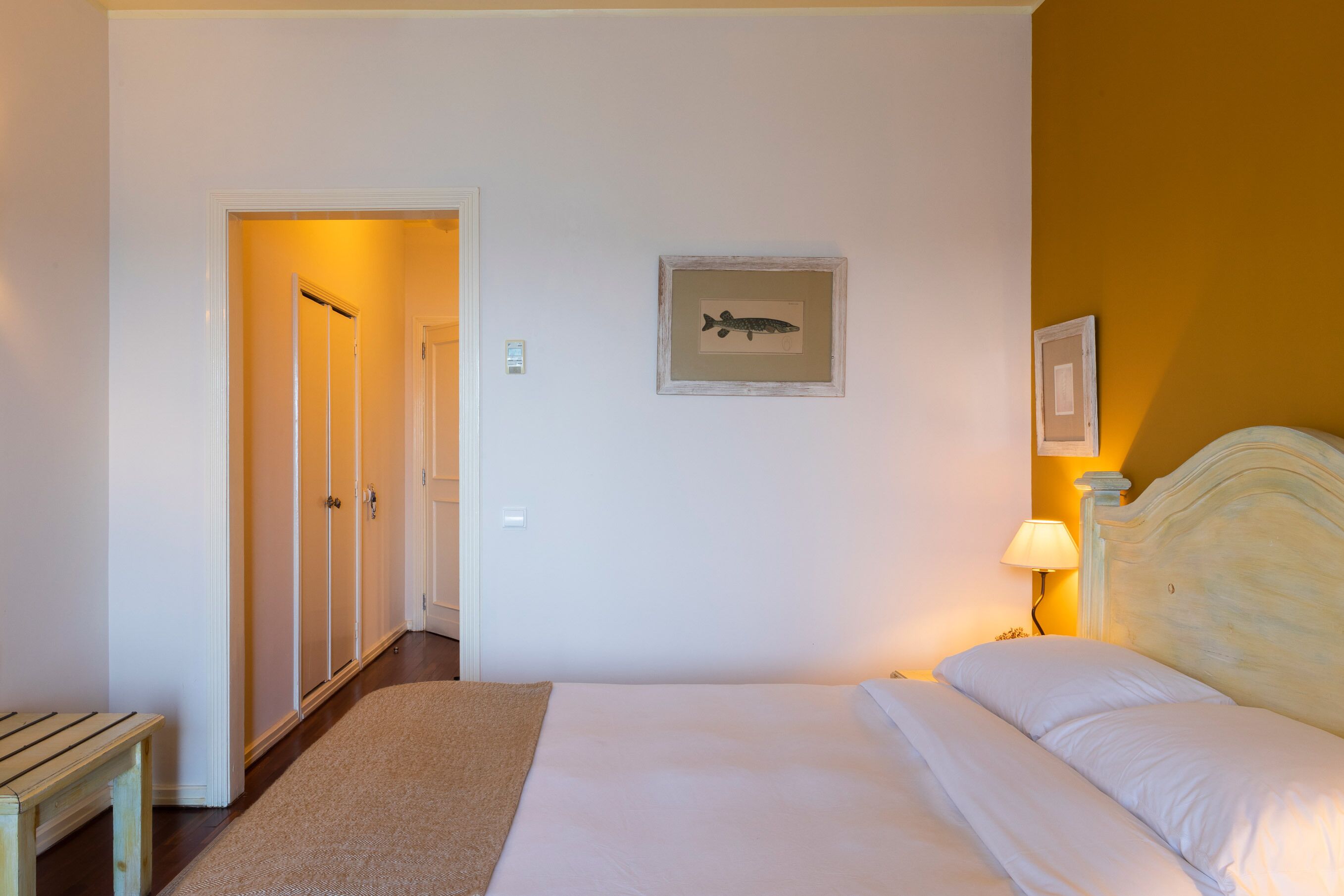 A hotel room with a bed, a bedside table, a lamp, a framed picture on the wall, and a door leading to another room.