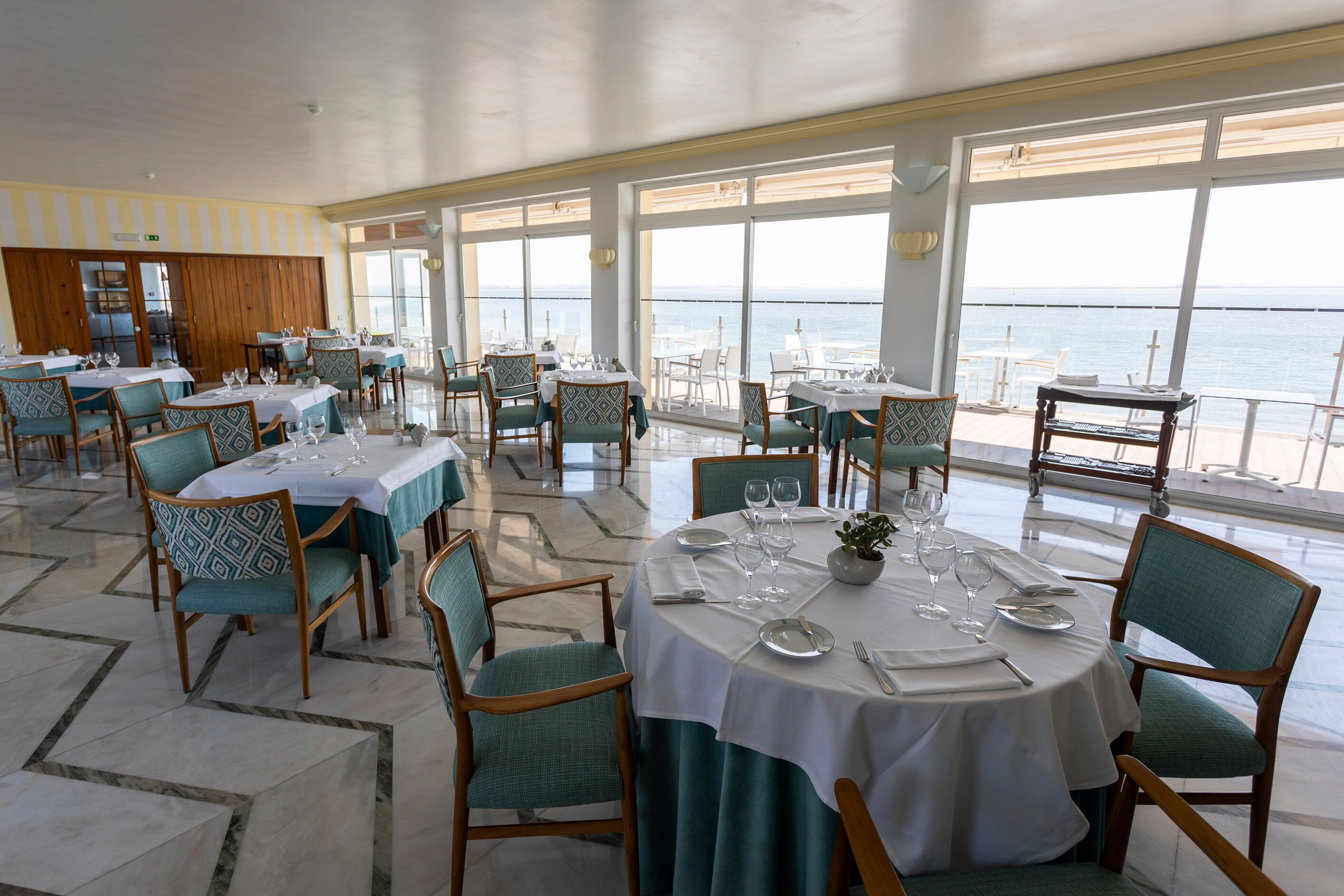 Un comedor con mesas preparadas para el servicio, amplias ventanas que ofrecen vista al mar y un panel de pared de madera al fondo.