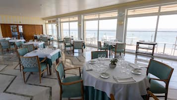 Un comedor con mesas preparadas para el servicio, amplias ventanas que ofrecen vista al mar y un panel de pared de madera al fondo.