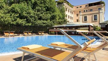 A swimming pool with sun loungers and a covered seating area, surrounded by trees and a building with balconies.