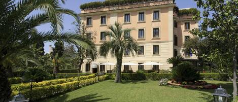 Un hotel de varios pisos con una fachada simétrica, un jardín cuidado con palmeras y setos, y área de estar al aire libre con sombrillas.