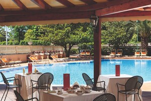 Spiseområde ved poolen med borde og stole, en pool og udsigt til frodigt grønt område.