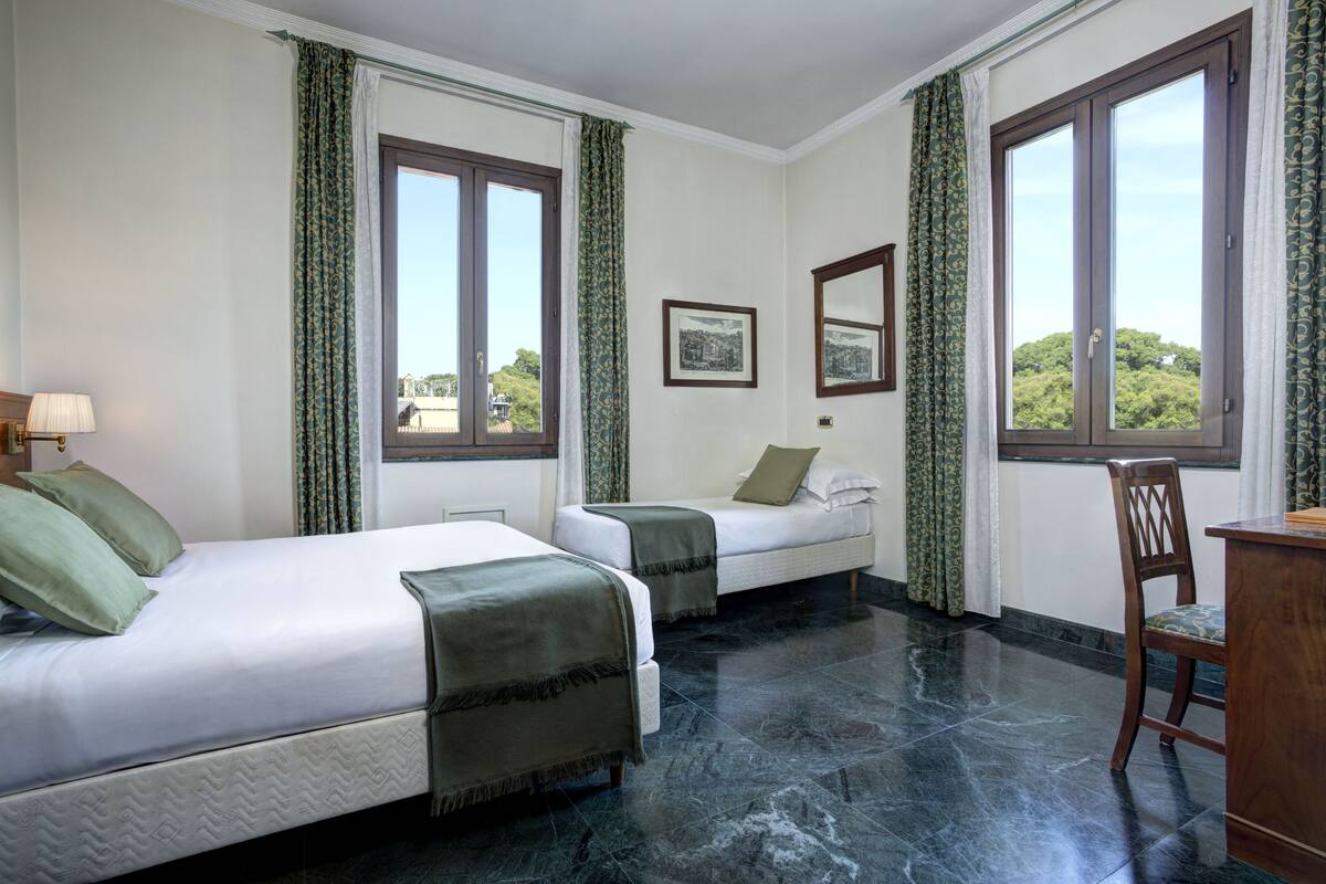 A hotel room with two beds, a desk, and a chair. There are large windows with curtains, and a picture on the wall.