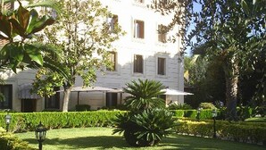 Un edificio blanco con numerosas ventanas y un jardín cuidado que incluye césped, setos y una palmera.