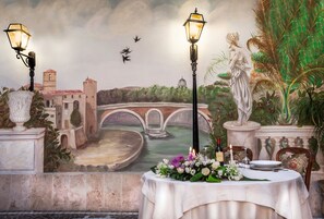 A dining table set with flowers and candles, a mural of a bridge and cityscape, and two street lamps.