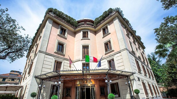 Un hotel histórico con una fachada clásica europea, una entrada central con puerta de vidrio y banderas en el tejado.