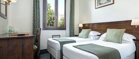 A hotel room with two single beds, a wooden dresser, a desk with a lamp, a window with curtains, and a framed picture on the wall.