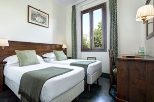 A hotel room with two beds, a wooden headboard, a window with curtains, a desk with a lamp, and a framed picture on the wall.