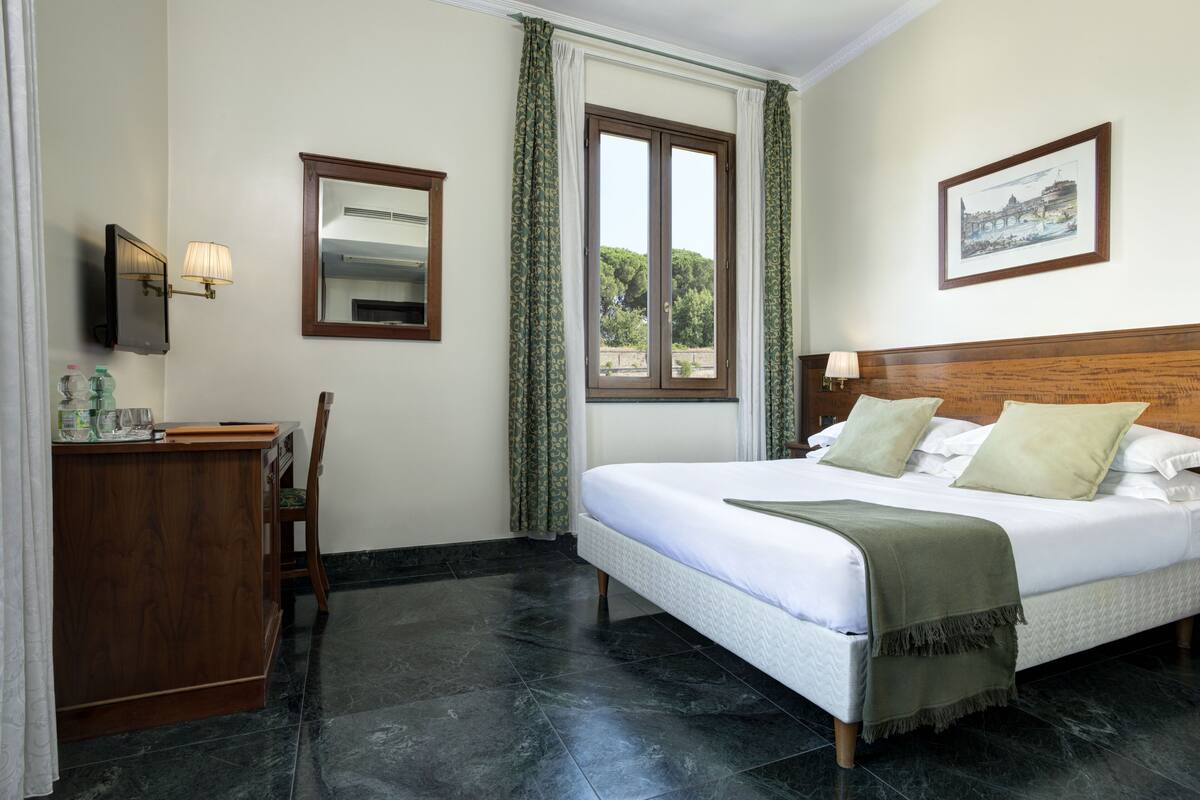 A hotel room with a bed, a desk, a chair, a window with curtains, a wall-mounted TV, and a framed picture.