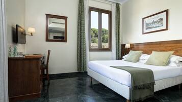 A hotel room with a bed, a desk, a chair, a window with curtains, a wall-mounted TV, and a framed picture.