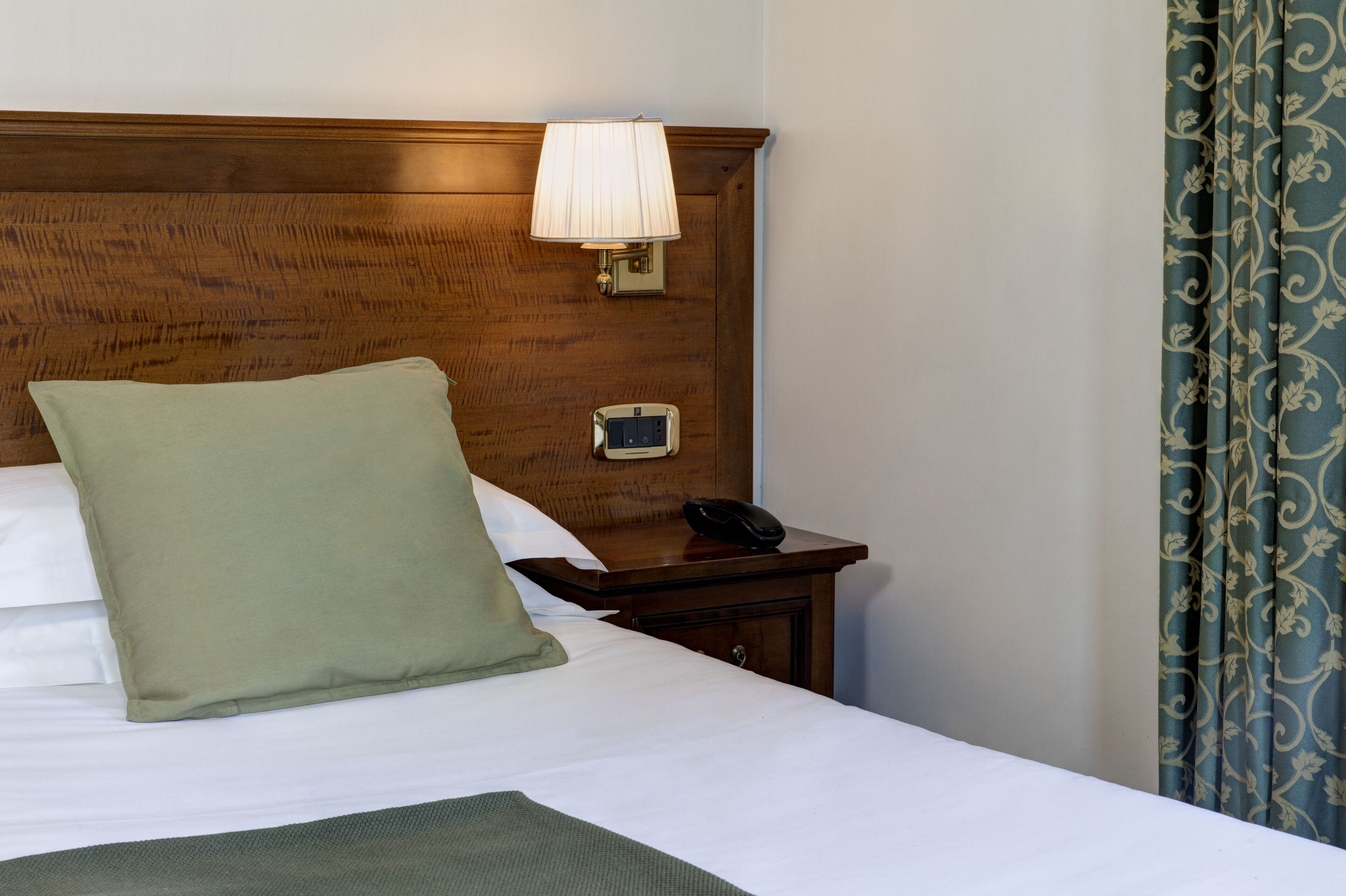 Una cama bien hecha con un cojín verde, una mesita de noche con una lámpara y un teléfono.