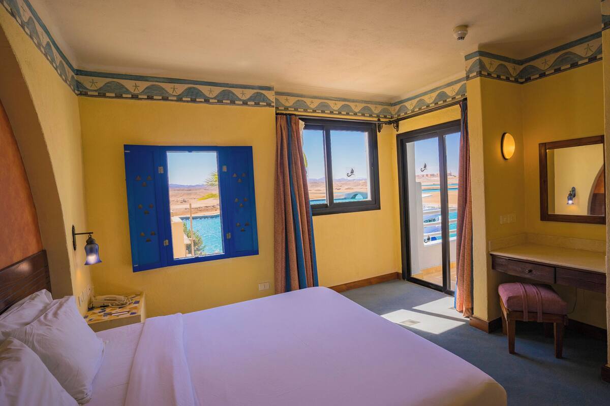 Ein Hotelzimmer mit Bett, Schreibtisch, Stuhl, einem Fenster mit Blick auf eine Wüstenlandschaft sowie einem Badezimmer mit Dusche und Toilette.