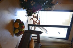 A room with a large window, a wooden chair, a table with a fruit bowl, and a decorative plant arrangement.