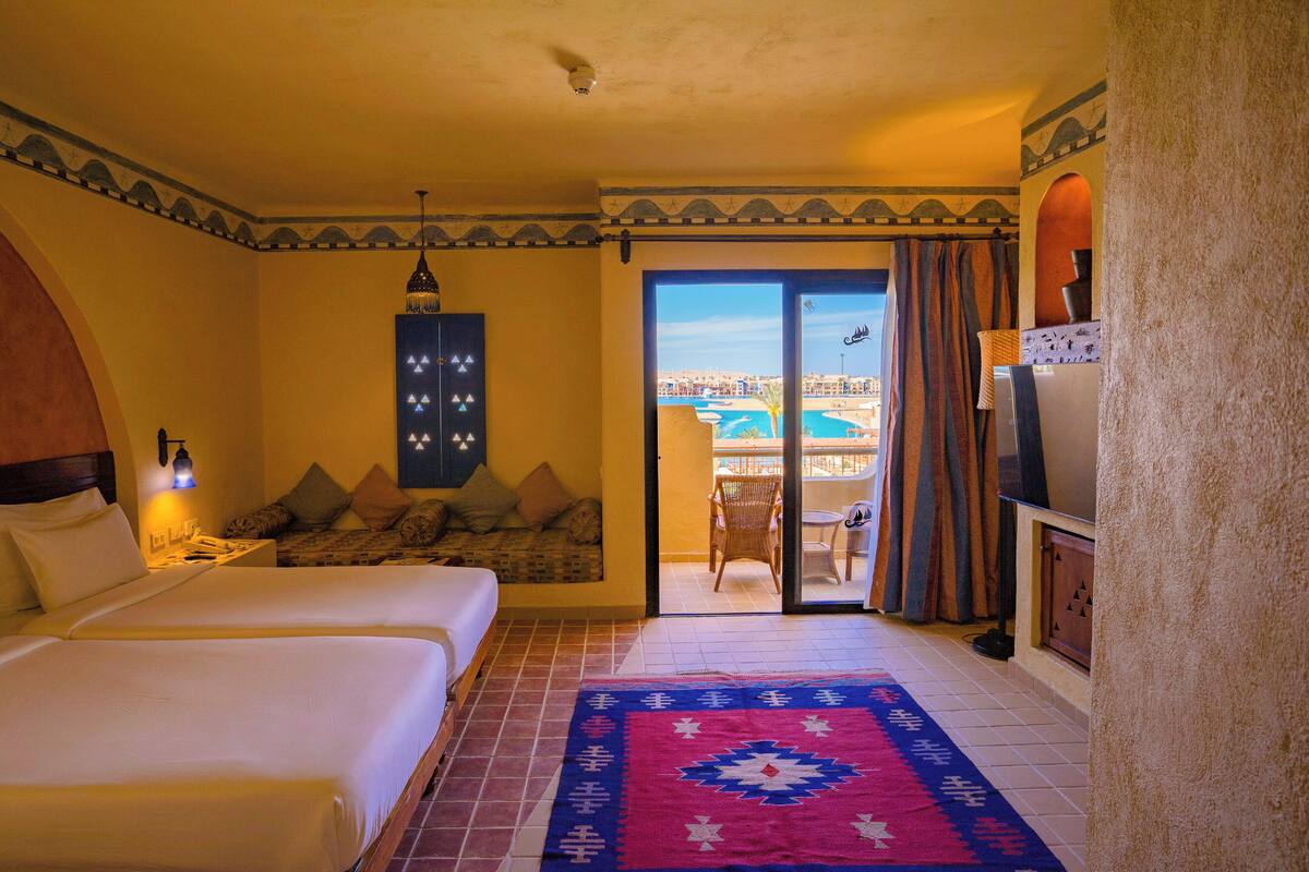 Ein Hotelzimmer mit zwei Betten, einem gemusterten Teppich, Blick auf einen Pool und Gebäude sowie einem Balkon mit Stuhl und Tisch.