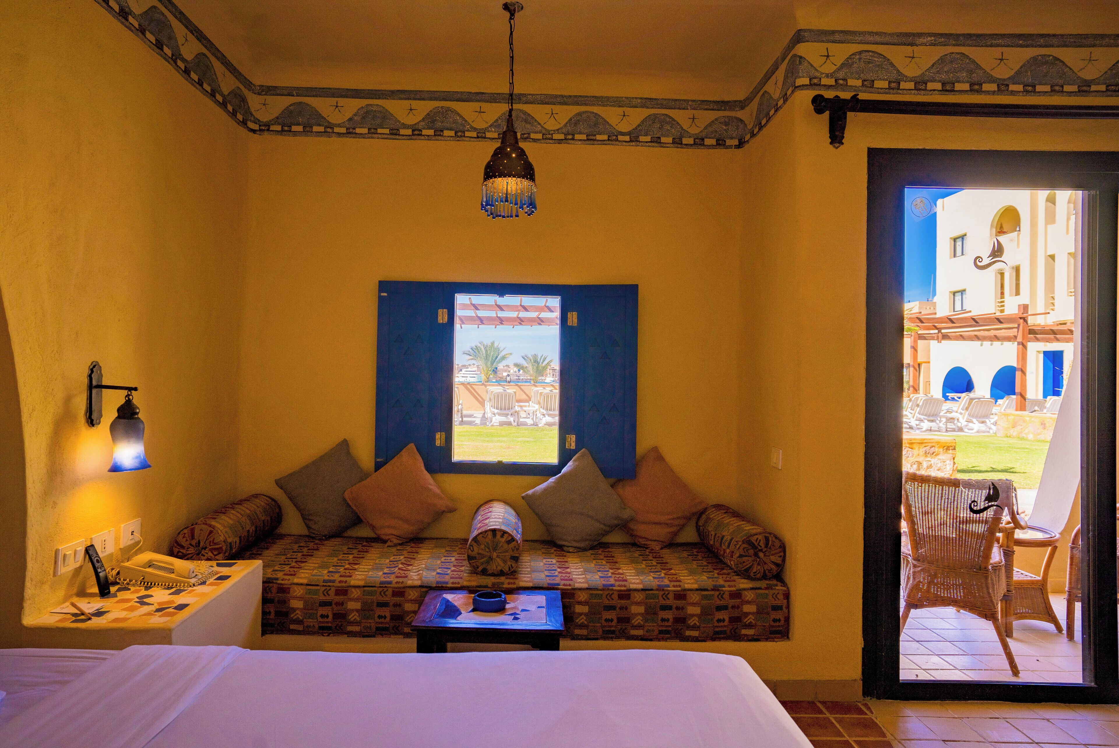 A room with a bed, a sofa with cushions, a small table, a lamp, and a view of the outside.