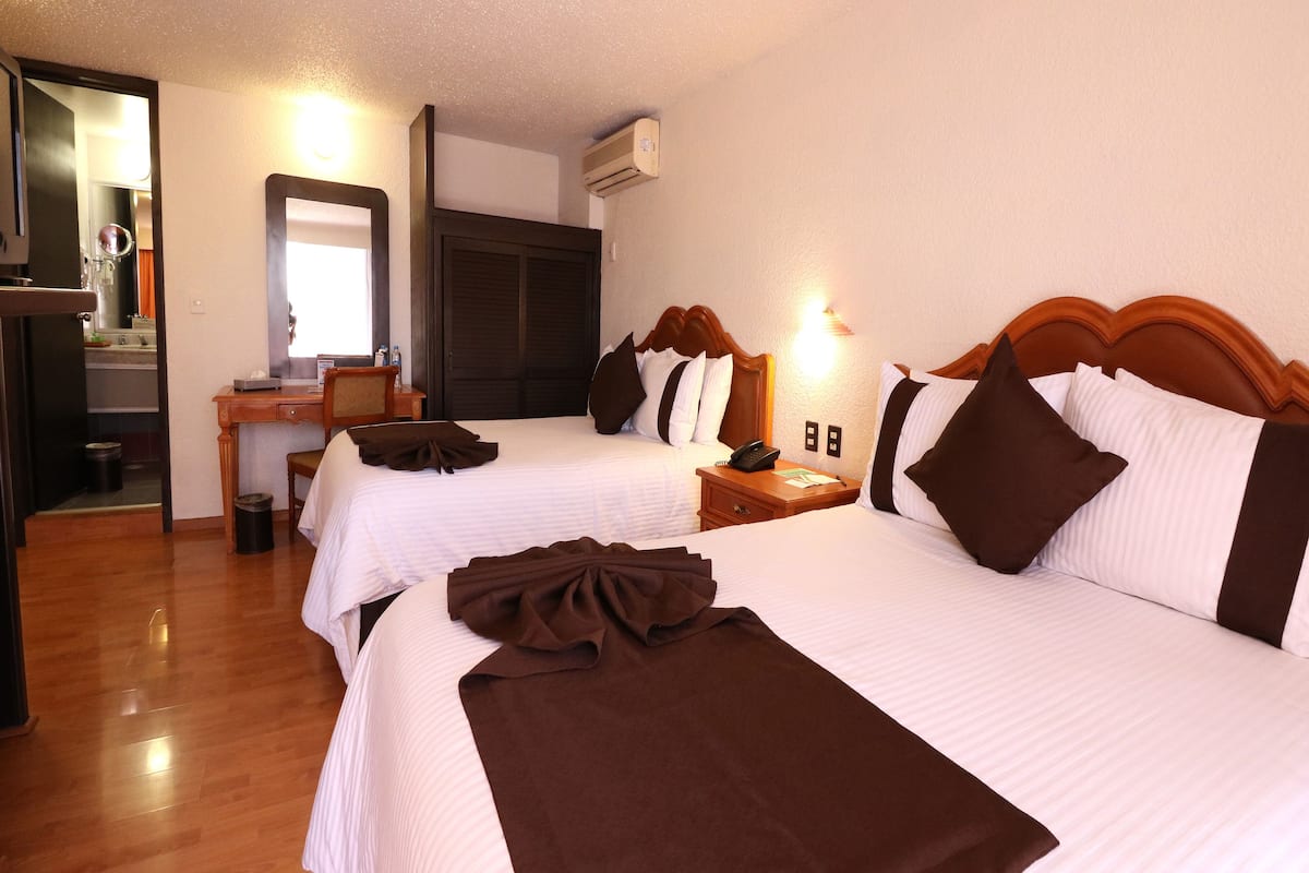 A hotel room with two beds, a desk, a chair, a mirror, and an air conditioning unit.