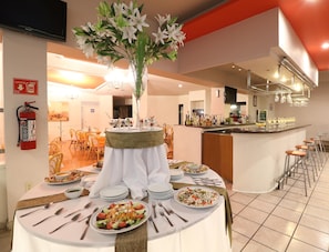 A dining area with a central table set for a meal, surrounded by chairs, a bar area with a counter and stools, and a fire extinguisher mounted on the wall. - Hotel Misión Aguascalientes Zona Sur (Aguascalientes)