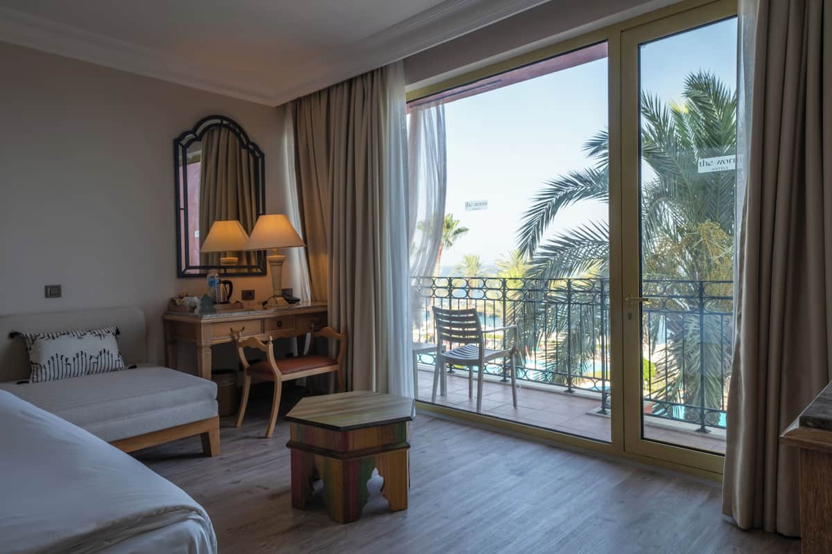 Habitación de hotel con cama, escritorio con lámpara, una mesita y balcón con vista a palmeras.
